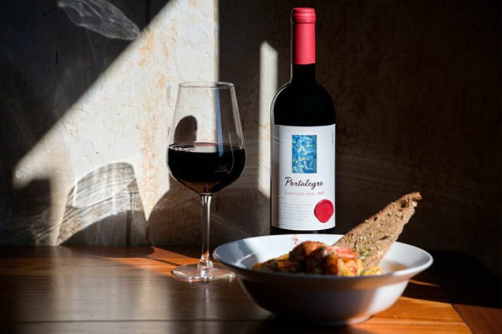 Weinflasche und Glas stehen auf Holztisch, neben Schüssel mit Essen, im Sonnenlicht.