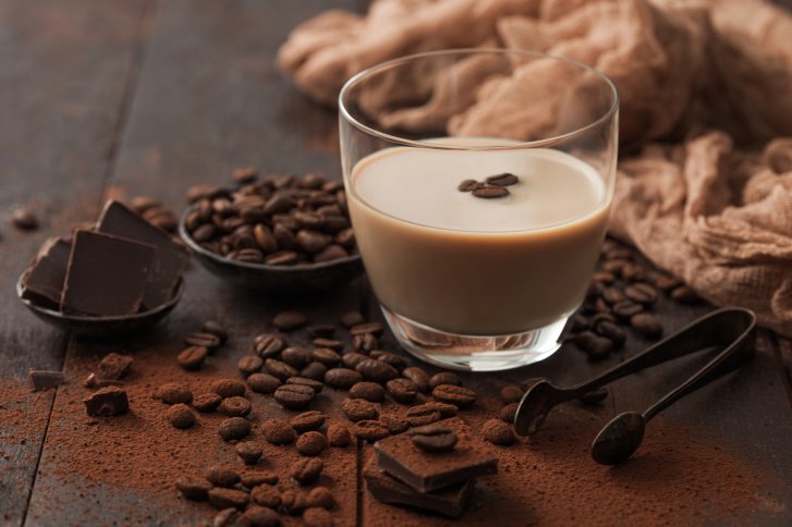 Ein Glas Sahnelikör steht auf einem Holztisch, umgeben von Kaffeebohnen und Schokolade.
