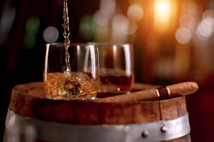 Whiskey wird in ein Glas auf einem Holzfass gegossen, daneben liegt eine Zigarre.
