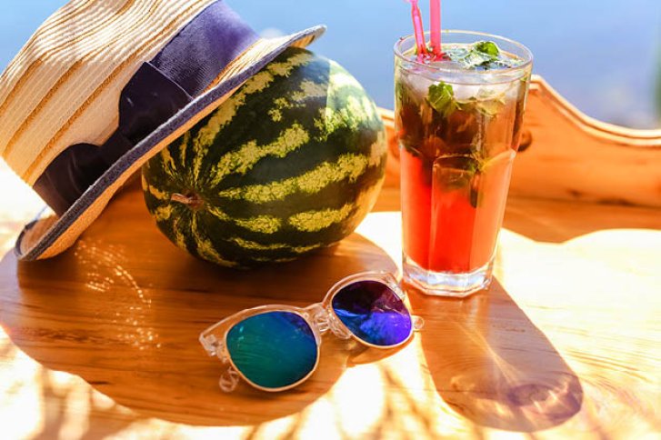 Hut, Wassermelone und Sonnenbrille liegen neben Cocktail auf sonnigem Holztisch.