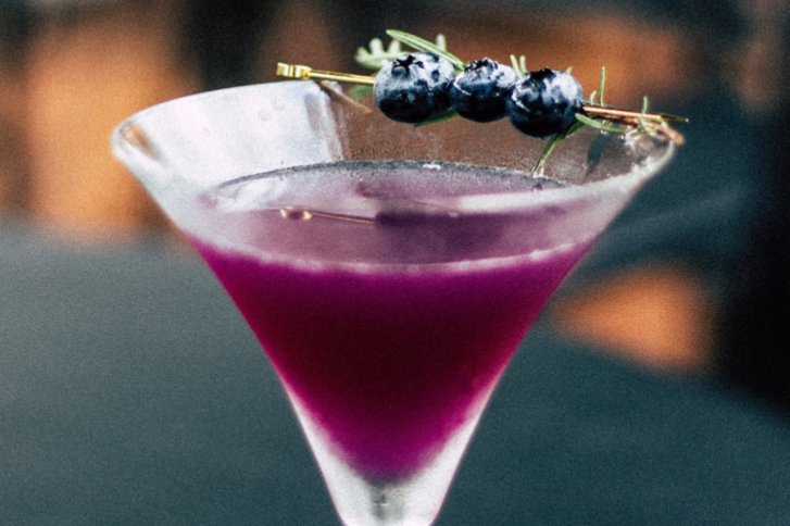 Lila Cocktailglas mit Blaubeeren garniert, steht auf einem Tisch in unscharfem Hintergrund.