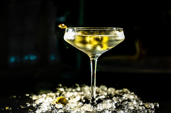 Cocktailglas mit Olive steht auf Eiswürfeln, dunkel beleuchteter Hintergrund kontrastiert die helle Flüssigkeit.