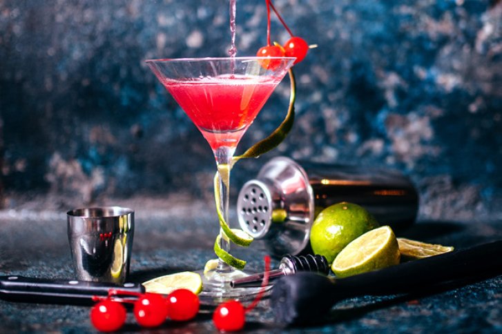 Cocktailglas mit rotem Getränk und Kirschen, umgeben von Limetten und Barzubehör vor dunklem Hintergrund.