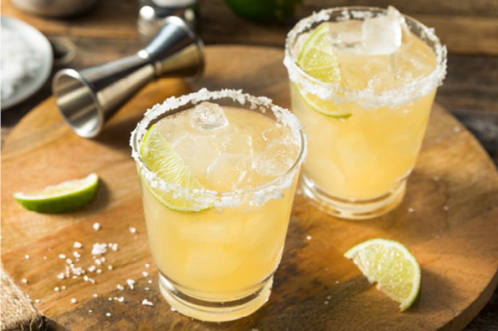 Zwei Gläser Margarita stehen mit Limettenscheiben und Eis auf einem Holzbrett, umgeben von Barzubehör.