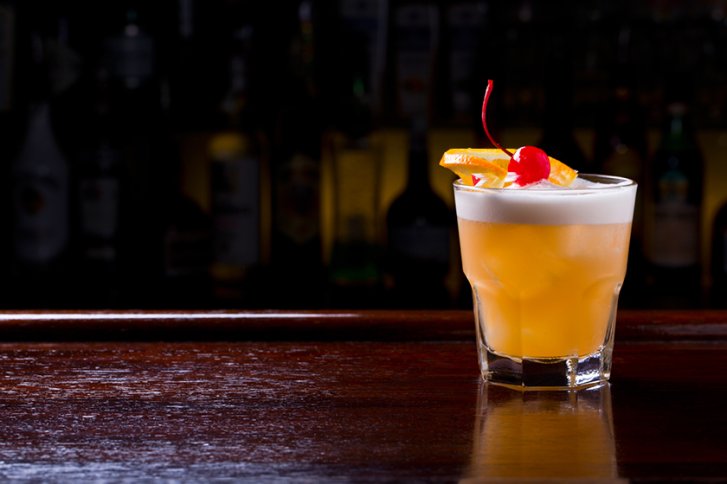 Ein Cocktailglas mit Schaum, dekoriert mit Kirsche und Orange, steht auf dunkler Bar.