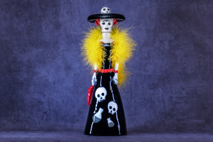 Skelettfigur trägt schwarzen Hut und gelbes Boa, gekleidet in schwarzem Kleid mit Totenköpfen.