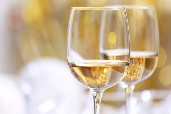 Zwei Weißweingläser stehen gefüllt vor einem verschwommenen, goldfarbenen, festlichen Hintergrund.
