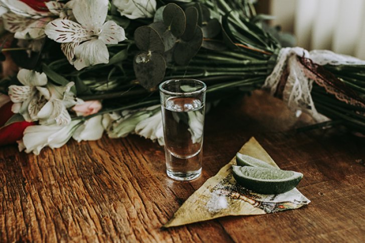 Ein gefülltes Schnapsglas steht neben Limettenscheiben und Blumenstrauß auf einem Holztisch.