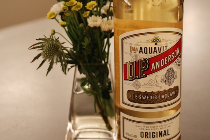 Flasche Aquavit steht neben Blumenstrauß in schlichtem, modernen Innenraum auf einem Tisch.