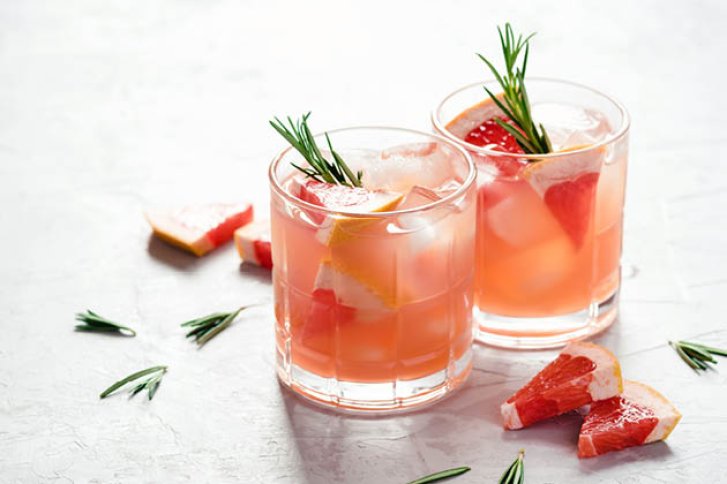 Zwei Gläser mit rosa Getränk, dekoriert mit Rosmarin und Grapefruit, stehen auf hellem Untergrund.