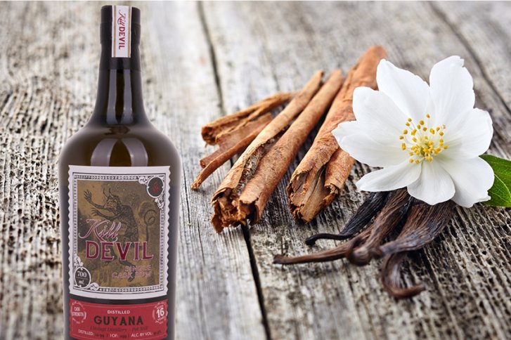 Rumflasche steht neben Zimtstangen, Vanilleschoten und einer Blume auf einer Holzoberfläche.