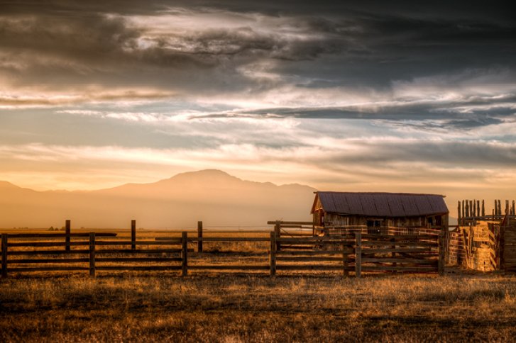 Holzscheune steht ruhig auf einem Feld bei Sonnenuntergang, umgeben von Bergen und Wolken.