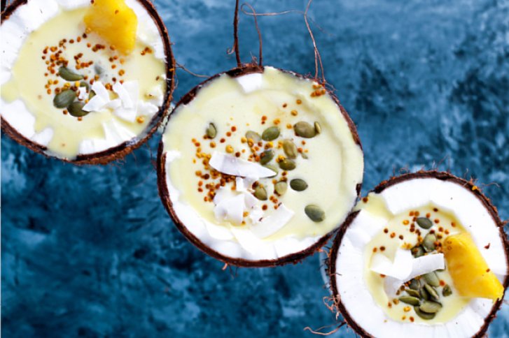 Drei Kokosnusshälften gefüllt mit gelber Creme, dekoriert mit Samen und Kokosflocken auf blauem Hintergrund.