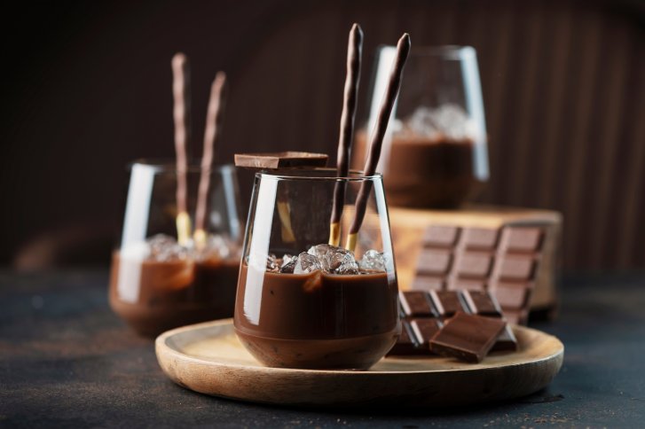 Gläser mit Schokoladengetränk, Eiswürfeln und Keksen, arrangiert neben Schokoladentafeln auf einem Holztablett.
