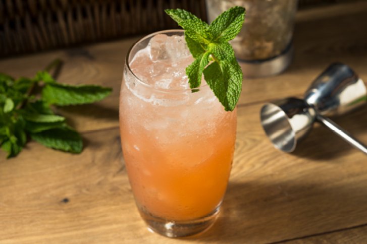 Ein Cocktailglas mit rosa Getränk und Minze darin steht auf einem Holztisch.