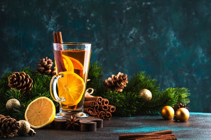 Glas mit Punsch, steht dekoriert mit Zimt und Orangen, zwischen Tannenzweigen und Zapfen.