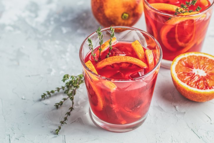 Rote Getränke mit Orangenscheiben und Thymian stehen auf einem hellen, strukturierten Tisch.