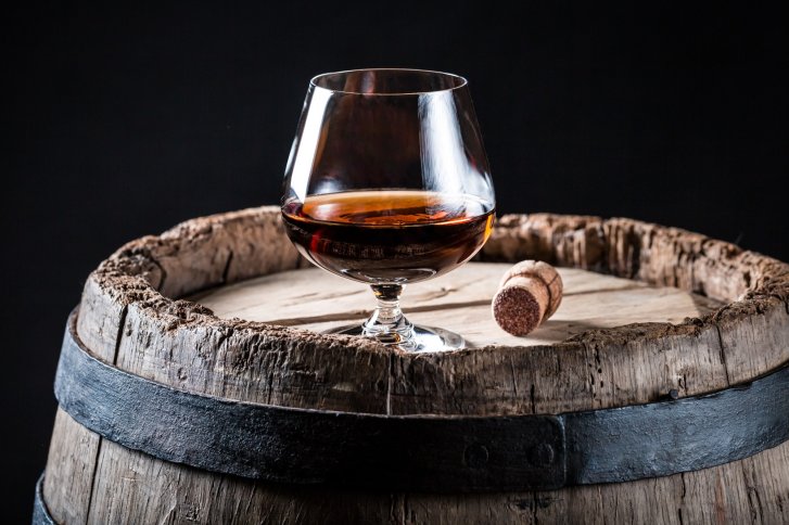 Ein Brandyglas steht auf einem Holzfass mit Korken, vor schwarzem Hintergrund.