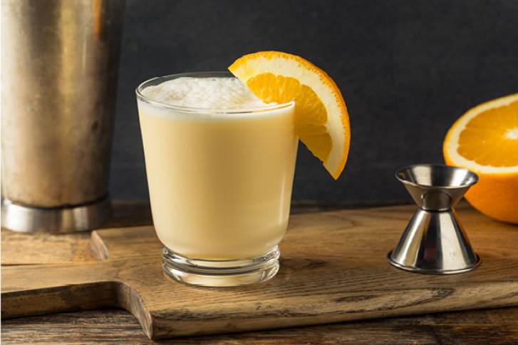 Ein milchiger Cocktail ruht auf einem Holzbrett, geschmückt mit einer Orangenscheibe.