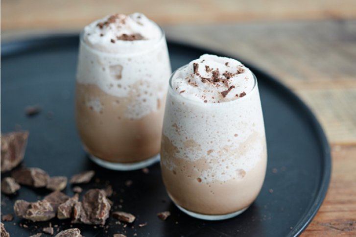 Zwei gläserne Becher mit Schokoladenshake, gekrönt mit Sahne und Schokoladenstücken, stehen auf einem Tablett.
