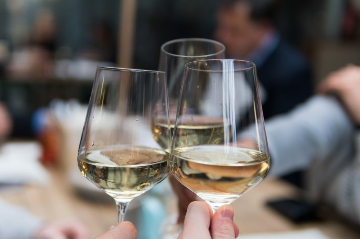 Drei Weingläser, gefüllt mit Weißwein, stoßen an, im Hintergrund unscharfe Menschen.