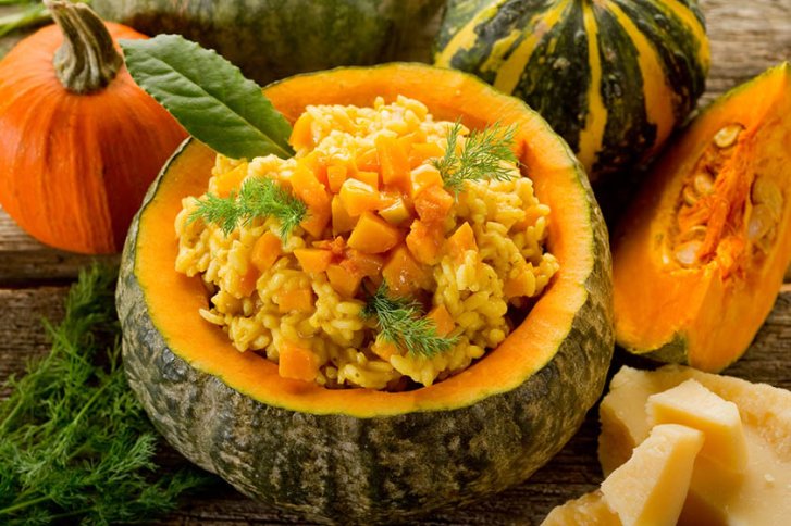 Runder Kürbis gefüllt mit Risotto steht auf rustikalem Holztisch, umgeben von Kürbisstücken und Parmesan.