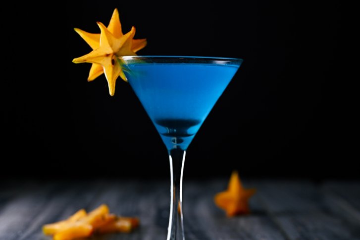 Blaues Cocktailglas mit sternförmiger Frucht, steht auf dunklem Holztisch vor schwarzem Hintergrund.
