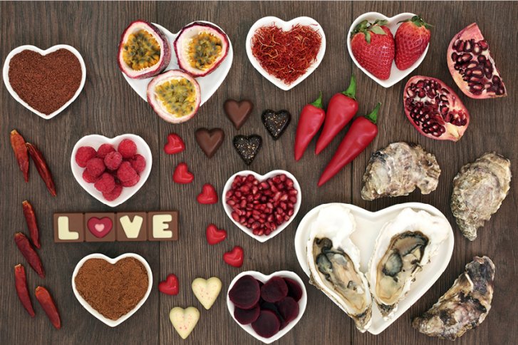 Herzförmige Schalen mit Früchten, Pralinen und Austern liegen dekorativ auf Holztisch.