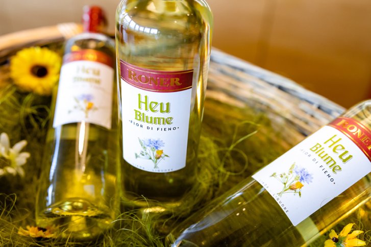 Flaschen mit "Heu Blume"-Etikett liegen auf Heu in einem Korb, dekoriert mit Blumen.