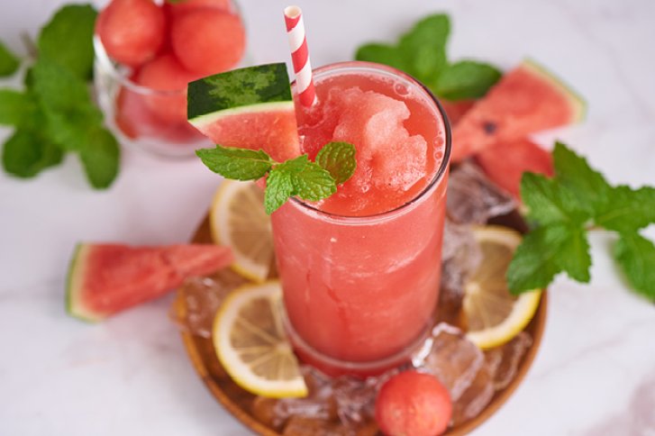 Ein rotes Wassermelonensmoothie-Getränk dekoriert mit Minze und Zitronenscheiben auf Eis in sommerlicher Umgebung.