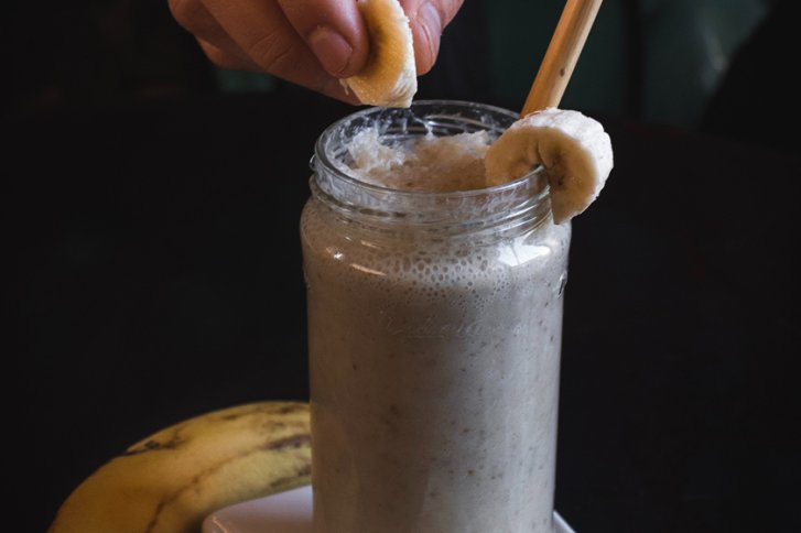 Ein Glas voller Smoothie, dekoriert mit Bananenscheiben, wird von einer Hand gehalten.