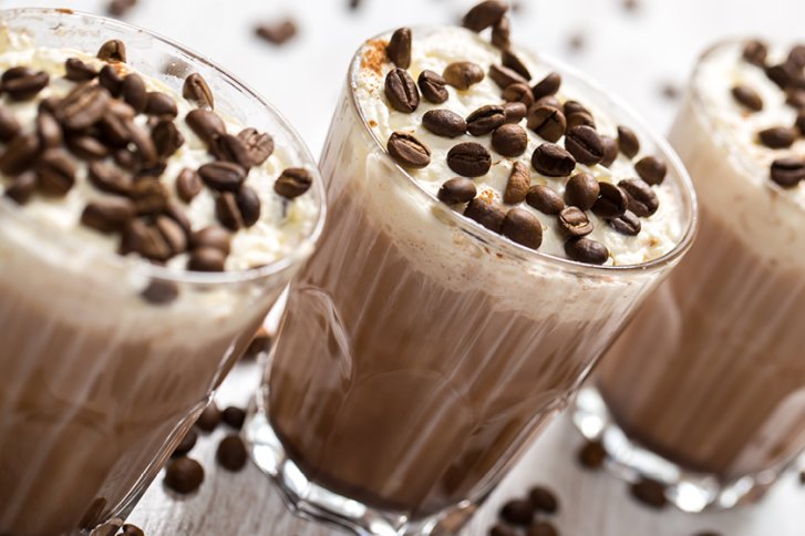 Drei Gläser gefüllt mit cremigem Kaffee, darüber geröstete Kaffeebohnen, auf einem Holztisch.