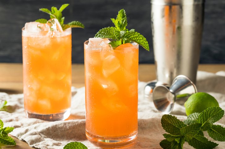 Zwei orange Cocktails stehen mit Eis und Minze garniert auf einem Tuch, umgeben von Zutaten.