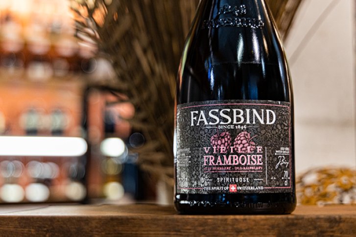 Eine schwarze Flasche steht auf einem Holztisch, Hintergrund unscharfe Regalbeleuchtung, Etikett "Fassbind Vieille Framboise".
