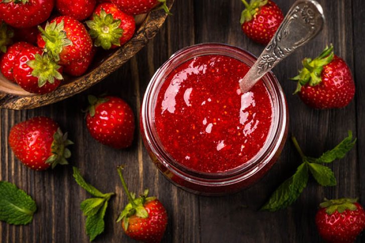 Ein Glas Erdbeermarmelade mit Löffel, umgeben von frischen Erdbeeren auf dunklem Holztisch.