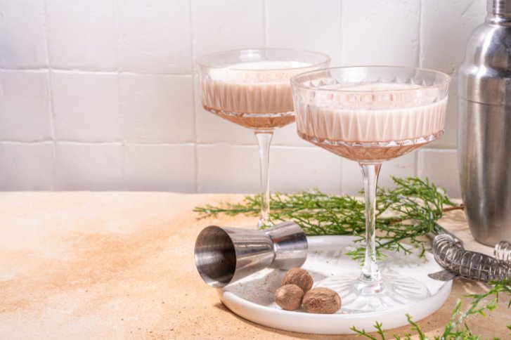 Brandy Alexander mit Shaker, zwei Gläsern, Portionierer, Muskatnuss und Zweigen auf hellem Hintergrund.