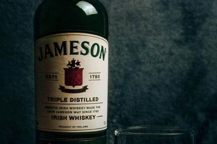 Eine Jameson-Whiskeyflasche steht auf einem Tisch, daneben ein leeres Glas, vor dunklem Hintergrund.