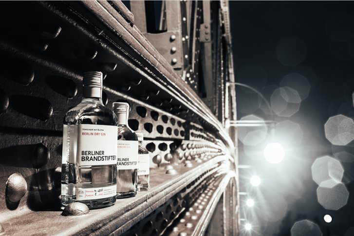 Zwei Ginflaschen stehen auf einer stählernen Brücke bei Nachtbeleuchtung. Text: "Berliner Brandstifter".