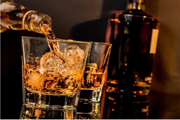 Glas wird mit goldenem Getränk aus Flasche gefüllt, im Hintergrund steht eine weitere Flasche.