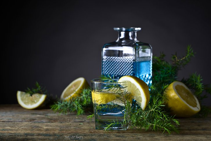 Glas Wasser mit Zitronenscheibe, vor einer Karaffe, umgeben von Wacholderzweigen auf Holztisch.