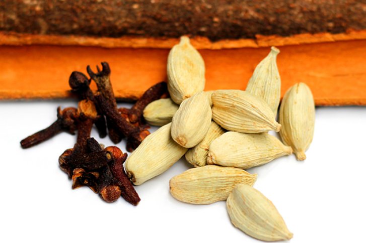 Gewürznelken und Kardamomkapseln liegen auf Zimtstange, vor weißem Hintergrund.
