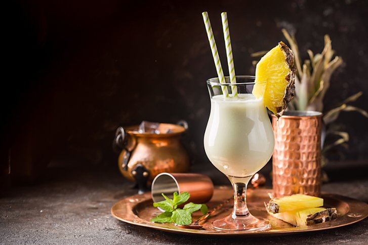 Ein Cocktailglas mit Ananasscheibe und Strohalmen steht auf Kupfertablett, umgeben von Dekoration.