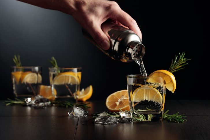 Hand gießt Getränk aus Shaker in Glas, umgeben von Zitronenscheiben und Rosmarin.