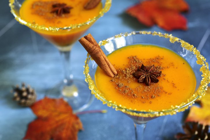 Zwei Cocktails mit orangener Flüssigkeit, Zimtstange, Sternanis in Martini-Gläsern auf herbstlichem Hintergrund.