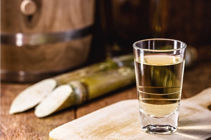 Ein Glas mit hellem Getränk steht auf Holzplatte, neben Zuckerrohrstücke und Fass.