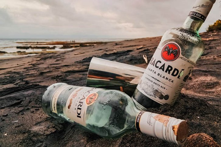 Zwei Bacardi-Flaschen liegen im Sand am Strand, daneben ein silberner Becher, Meer im Hintergrund.