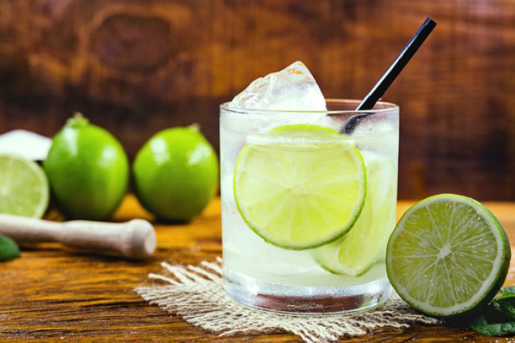 Ein Glas Caipirinha mit Limettenscheiben und Eiswürfeln steht auf Holztisch, umgeben von Limetten.