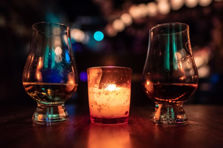 Zwei Gläser mit Getränken stehen neben einer brennenden Kerze auf einem Tisch in dunkler Atmosphäre.