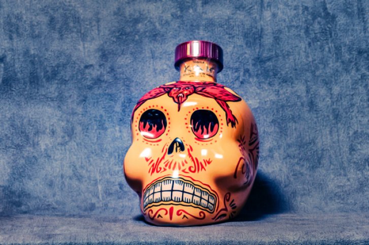 Schädel-Flasche symbolisch verziert, steht auf blauer Oberfläche, Hintergrund ebenfalls blau strukturiert.