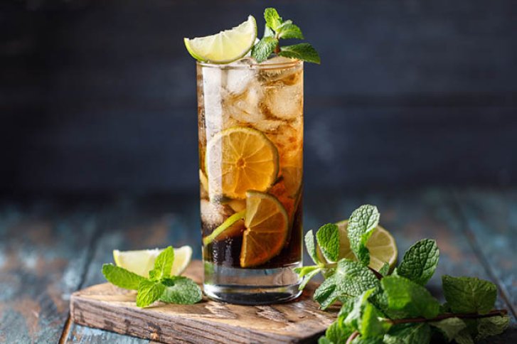 Ein hoher Cocktail mit Eiswürfeln, Limetten, Minzblättern auf einem rustikalen Holztisch arrangiert.